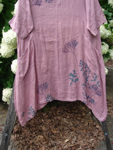 Bluefishfinder.com Barclay Linen Wave Pocket Dress in dried sprig tulsi rose, size 1, a loose pink-lavender linen dress with a softly crinkled texture, short sleeves, and a gently curved hem. Blue floral sprig prints cluster near the lower front as it hangs against leafy white blooms.