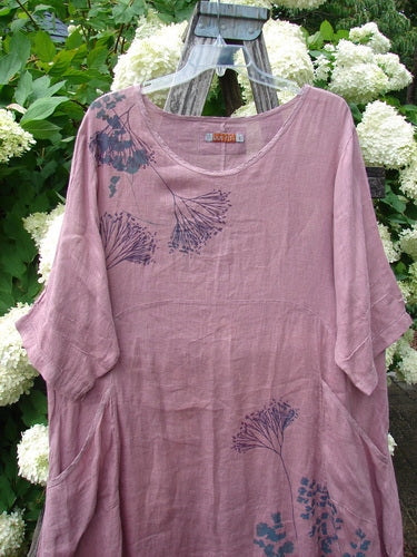 Bluefishfinder.com Barclay Linen Wave Pocket Dress in Dried Sprig Tulsi Rose, size 1, a loose mauve linen dress with a soft washed texture and dark botanical print. It hangs on a wooden hanger, its wide neckline and relaxed sleeves framed by white hydrangeas and green foliage.