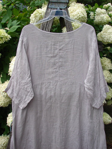 Bluefishfinder.com Barclay Linen Wave Pocket Dress in Fern Rice Paper, size 1, shown in a soft gray linen with a loose, draped silhouette and wide boat neckline. Short sleeves, subtle front seam detail, and a lightly textured finish are visible against leafy white hydrangeas.