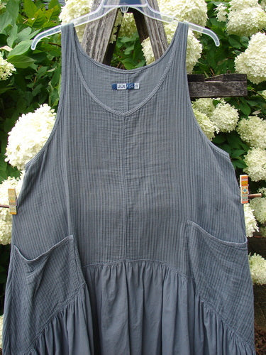 Bluefishfinder.com Barclay Gauze Batiste Summer Frock Jumper in unpainted gray day stripe, a sleeveless dress with a scooped neckline, fine vertical texture, side pockets, and gathered skirt. The lightweight gauze batiste hangs on a hanger against white hydrangeas and dark wood.