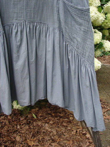 Bluefishfinder.com Barclay Gauze Batiste Summer Frock Jumper in unpainted gray day stripe, shown in a close-up of the skirt and side panel with soft gauze batiste texture, gathered seams, and a flowing, uneven hem. The muted gray fabric hangs against outdoor greenery and leaf litter.
