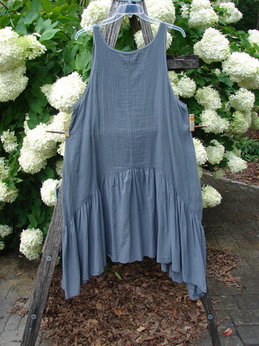 Bluefishfinder.com Barclay Gauze Batiste Summer Frock Jumper in unpainted gray day stripe, a sleeveless dress with a softly gathered empire seam and a tiered, fluttering hem. The gauzy batiste fabric hangs from a clear hanger, its fine striped texture set against white hydrangeas and weathered wood.