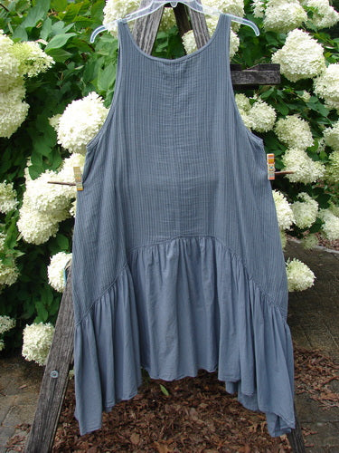 Bluefishfinder.com Barclay Gauze Batiste Summer Frock Jumper in unpainted gray day stripe, a sleeveless gauzy dress with a softly gathered yoke, vertical texture, and a fuller ruffled hem. It hangs from a wooden ladder outdoors, framed by white blossoms and green leaves.