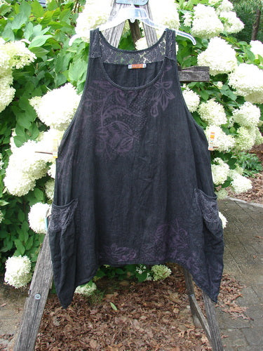 Bluefishfinder.com Barclay Summercloth Lace Two Pocket Pinafore in Moody Night Bloom Black, size 0, shown as a loose black pinafore with a soft lace-like floral pattern and two side pockets. The lightweight fabric hangs in a relaxed A-line shape against bright white blossoms and green foliage.