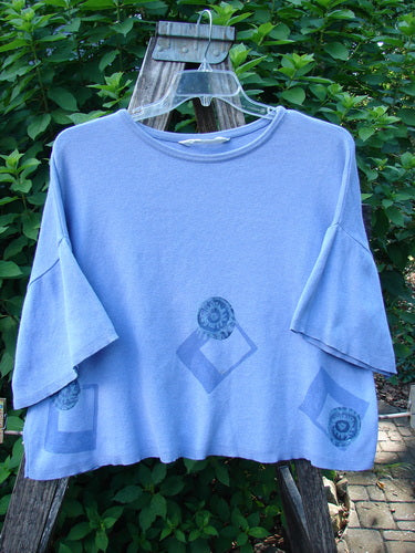 1997 Cotton Cashmere Short Sleeved Crop Pullover Geometric Skylark OSFA with wide scooped neckline, flared sleeves, and blue geometric motifs, displayed on a hanger outdoors against lush foliage.