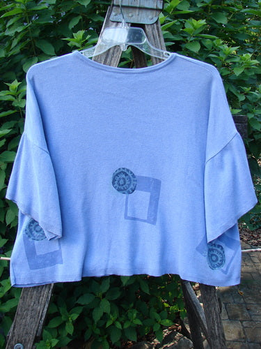 1997 Cotton Cashmere Short Sleeved Crop Pullover Geometric Skylark OSFA hangs on a clear plastic hanger, showcasing wide elbow-length sleeves and geometric screen-prints, set against lush green foliage.