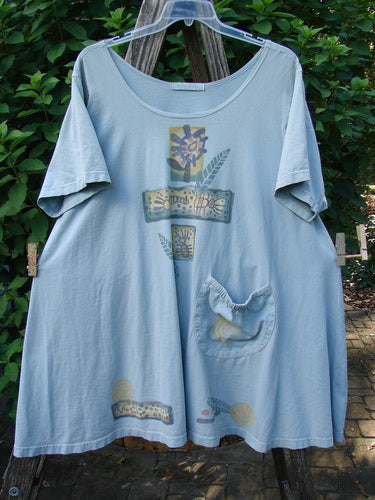 1995 Hop Scotch Dress Garden Friends Beach Glass Size 2, featuring a loose, light-blue tunic with abstract prints and a patch pocket, elegantly draped on an outdoor easel, showcasing vintage Blue Fish Clothing's unique style.