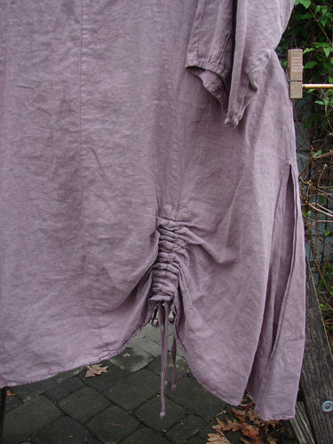 Barclay Linen High Vent Venetian Dress Celtic Fern Lavender Size 1 hangs elegantly on a clothesline, showcasing its draped fabric, softly dipped V neckline, and distinct curved seams.