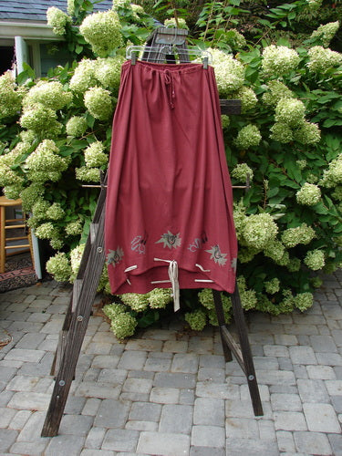 1992 Buttonloop Skirt Pomegranate Music OSFA displayed on a wooden rack, showcasing its full drawcord waist, slight bell shape, varying hemline, and button holes with hand-dyed silk ribbon.