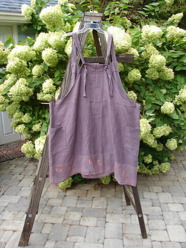 Barclay Linen Patterson Flutter Apron Jumper Butterfly Dusty Plum Size 2 displayed on a wooden rack, featuring mega sectional panels, batiste flutter hem, wrap exterior side pockets, and adjustable shoulder straps.