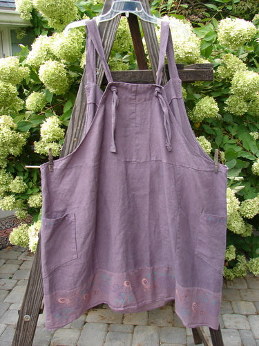 Barclay Linen Patterson Flutter Apron Jumper Butterfly Dusty Plum Size 2 displayed on a wooden stand, showcasing its adjustable shoulder straps, mega sectional panels, and exterior side pockets.