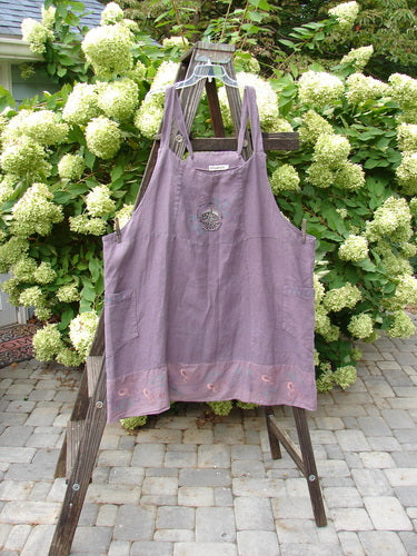 Barclay Linen Patterson Flutter Apron Jumper Butterfly Dusty Plum Size 2: A purple apron with butterfly-themed batiste flutter and wrap pockets on a wooden stand.
