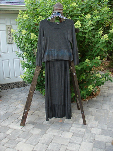 2000 Rayon Lycra Midi Bubble Trio Sparkle Border Charcoal Size 1 2: Clothes on a rack, including a long grey shirt and skirt on a wooden ladder, a white door with a sign, a black curtain with a black cloth, and a pair of glasses on a wooden stand.