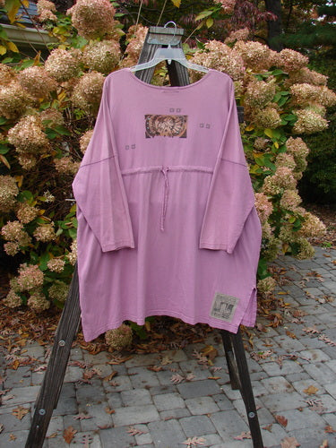 1997 Collage Top Petal Power Crocus OSFA: A pink shirt on a rack, featuring a long-sleeved purple shirt. Made from mid-weight organic cotton, it has an A-lined shape, drop shoulders, vented sides, and a varying hemline. Accented by vintage knot buttons, it has oversized front pockets and a nifty drawcord back. The superior Blue Fish Petal Power theme and the traditional Blue Fish Patch complete the look. Bust 66, waist 66, hips 68, front length 30, back length 35 inches.