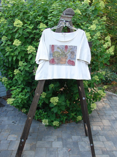 Barclay Crop Tee Top Bountiful Cacti Natural Altered Size 1 displayed on a wooden easel, showcasing its generous sleeves, wider neckline, and a vibrant cacti-themed design.