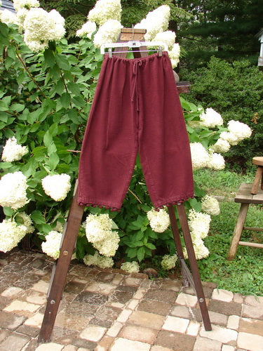 Image alt text: 1999 Flannel PJ Pant Pom Pom Unpainted Wine Size 0 - A pair of pants on a rack with a wooden bench and green leaves in the background.