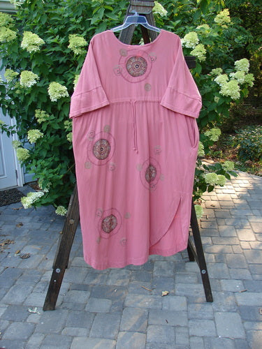Barclay Naiad Dress Medallion Glow Size 2: A pink dress on a clothes rack, featuring a super tapered lower shape, generous bust measurement, rear draw cord, shorter sectional lower sleeves, and a super rounded and vented hemline. Includes two deep side pockets and the dynamic Barclay Medallion Theme Paint. Made from mid-weight organic cotton. Length is 54 inches.