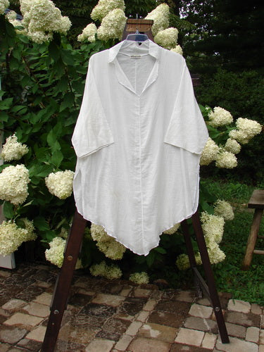 Image alt text: Barclay Batiste Flutter Tunic Top, a white shirt on a rack, featuring a V neckline, flutter collar, and varying front and back hemline.