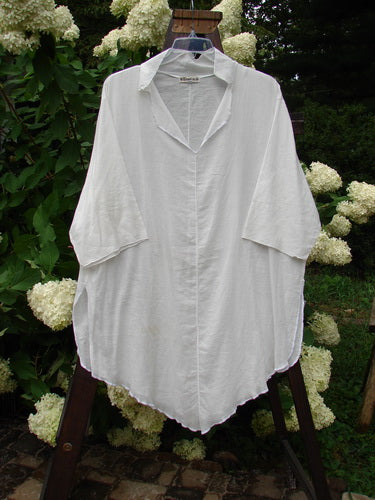 Barclay Batiste Flutter Tunic Top in Unpainted White, displayed on a rack, showcasing its V neckline, folded collar, flutter rear collar, and three-quarter length sleeves with soft exterior stitchery.