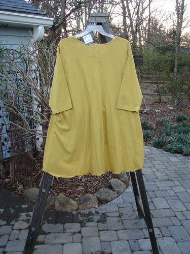 Barclay NWT High Low Top in Dusty Yellow, displayed on a clothes rack, showcasing its A-line shape, rounded banded bottom, thinner neckline, and varying hemline for a stylish, organic cotton look.