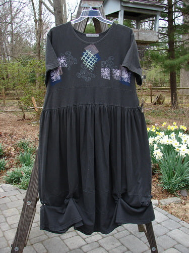 Black 1996 Constellation Dress with adjustable hemline, garden path theme paint, and buttons on hem and rear waistline, displayed on a wooden ladder. Size 2, organic cotton.