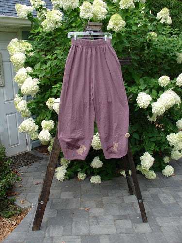 Image: A close-up of a pair of 1996 Boulevard Pants on a rack. The pants are made from organic cotton and have a unique old car travel theme paint design. The pants feature a two-inch elastic waistline, side seam pockets, and lower overlapping painted cuffs with cloth covered buttons. The measurements are: waist fully relaxed 26, waist fully extended 40, hips 50, inseam 23, and length 38 inches.