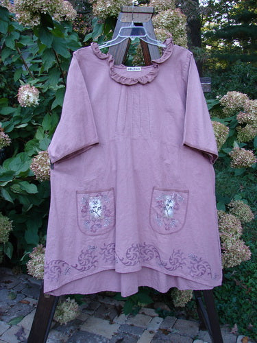 Barclay Linen Duet Sunrise Dress Tiny Floral Dusty Rose Size 2, featuring a ruffled neckline, pintuck bodice detail, elbow-length sleeves, floral patch pockets, and decorative hem, displayed on a wooden easel outdoors.