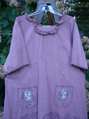 Barclay Linen Duet Sunrise Dress Tiny Floral Dusty Rose Size 2, featuring a ruffled neckline, elbow sleeves, and floral-embroidered pockets, hanging against green foliage, highlighting its loose fit and pin-tuck detailing.