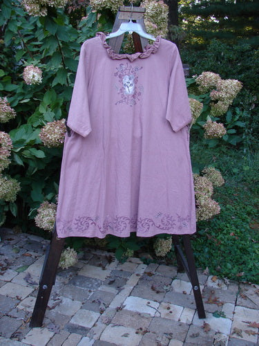 Barclay Linen Duet Sunrise Dress in Dusty Rose, featuring a ruffled neckline, elbow-length sleeves, and central floral motif, displayed outdoors on a wooden stand.