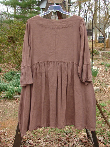 A brown dress on a clothes rack, part of the Barclay Linen Adras Uptown Jacket collection. Features include a deep V-shaped neckline, sectional triangular panels, pleated and banded lower sleeves and hemline, double exterior drop front pockets, and a varying hemline. Size 0, unpainted in redwood.