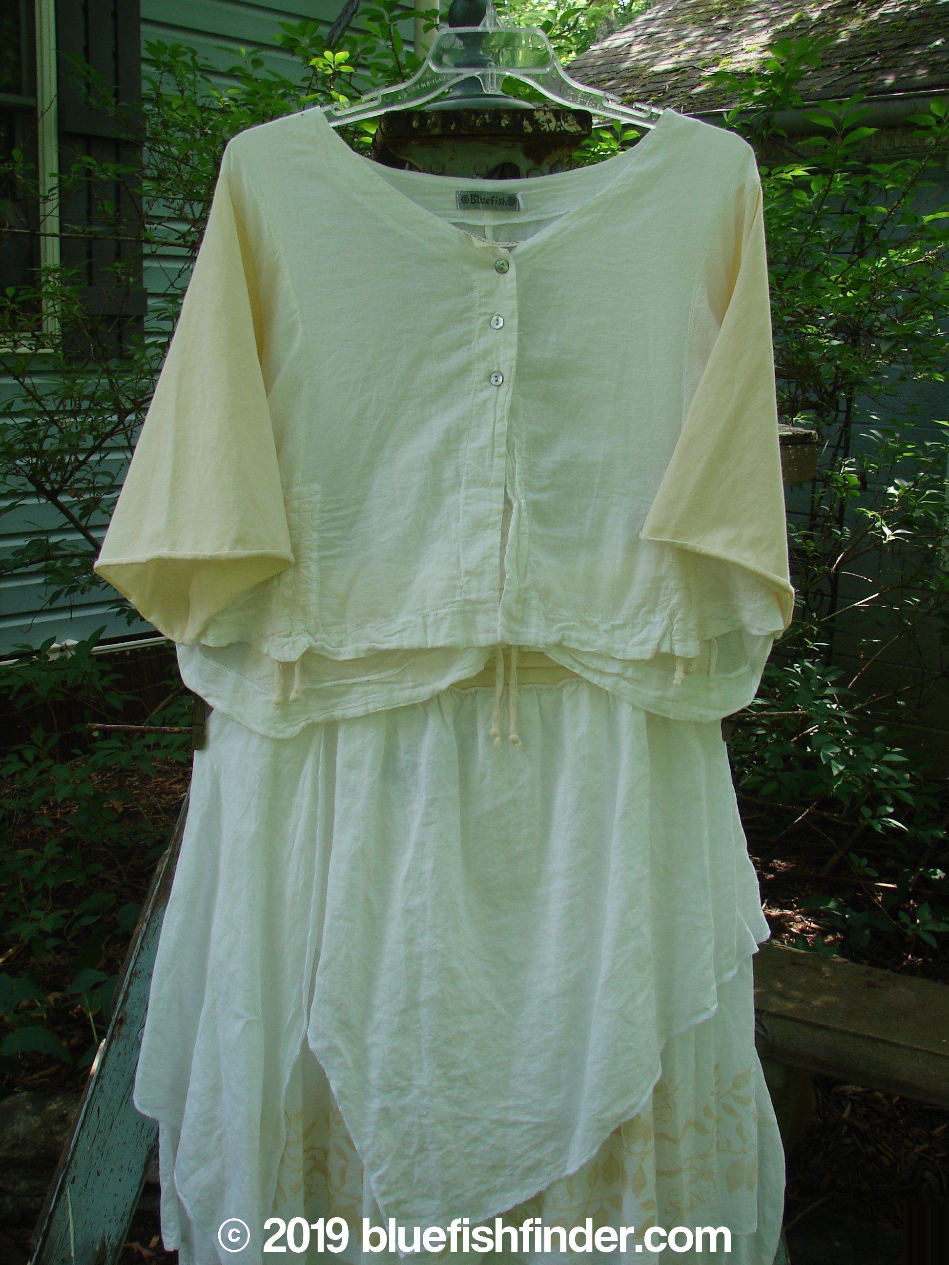 A white shirt and skirt on a clothesline, part of the Barclay NWT Batiste Draw Midi Meadow Trio in White. The shirt features a three-button front, three-quarter length sleeves, and a wider, boxier crop shape. The skirt has a full cotton paneled waistline, an outer layer of painted batiste set on the diagonal, and a sweeping batiste ruffled under layer. The set is made from a combination of organic cotton and batiste. Perfect for spring or summer!