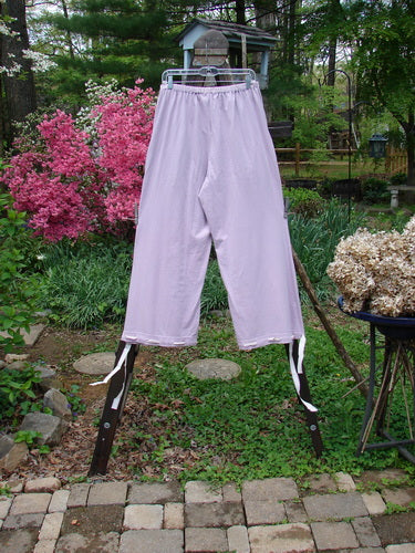 1999 Ribbon Pant Unpainted Orchid Size 1: A pair of pants on a rack, featuring a full elastic waistline, deep front bubble cargo pockets, and darling button holes on each cuff with silk ribbon. Playful and flared, these pants are perfect for a fun, fish-inspired look.