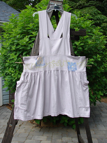 Image description: A vintage lavender overall jumper from the 1995 Spring Collection. The jumper features a creatively cut overall bib, a shorter length, a full gathered and wide playful skirt, two oversized side pockets, and crisscross back straps. It has a 6-inch double-stitched waistline panel, curly daisy theme paint, and the signature Blue Fish patch. Made from mid-weight organic cotton.