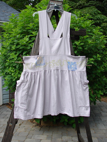 1995 Overall Jumper Daisy Curl Lavender OSFA displayed on a wooden fence, featuring an overall bib, crisscross back straps, oversized side pockets, and a gathered skirt in mid-weight organic cotton.