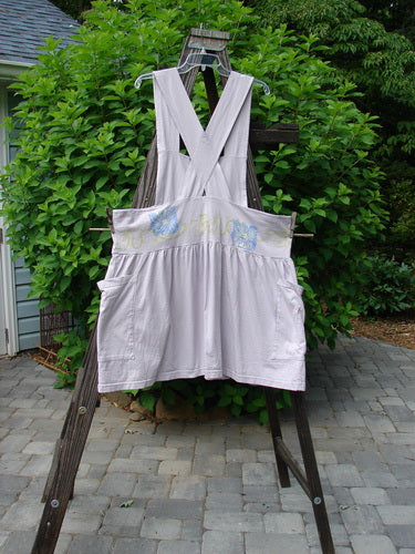 1995 Overall Jumper Daisy Curl Lavender OSFA displayed on a wooden rack, featuring a creatively cut bib, wide playful skirt, crisscross back straps, and oversized side pockets.
