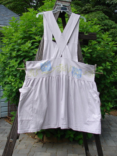 1995 Overall Jumper Daisy Curl Lavender OSFA displayed on a wooden structure, featuring a creatively cut bib, crisscross back straps, a gathered skirt, and two oversized side pockets.