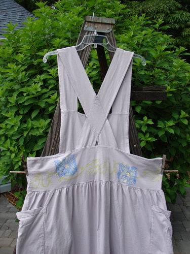 1995 Overall Jumper Daisy Curl Lavender OSFA displayed on a wooden rack, featuring a creatively cut bib, crisscross back straps, and a full gathered skirt with oversized pockets.