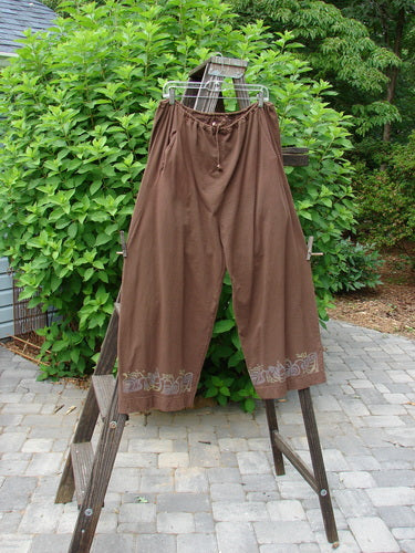 Image alt text: "1992 Buttonloop Pant Grape Leaf Bay Leaf OSFA: Brown pants with a full drawstring waistline, deep side pockets, and grape leaf paint, displayed on a wooden ladder."