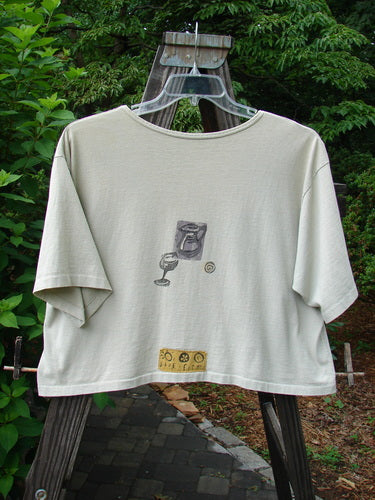 1991 Short Sleeved Oversized Tee Pitcher Glass Wheat OSFA displayed on a hanger, showcasing a vintage goblet theme drawing, wide boxy cut, rolled neckline, and blue fish patch, highlighting its collectible nature.