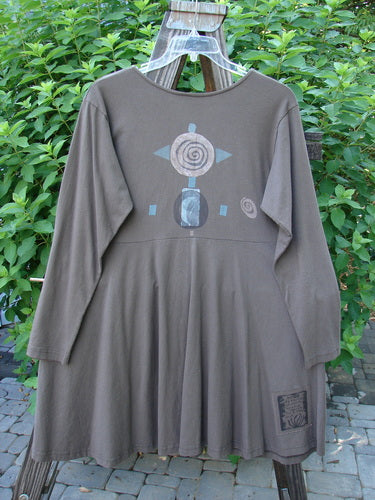 1994 Poppy Dress, collarless, in Humus, size 2. Long-sleeved dress on a clothes rack. Grey dress with a design. Drawing of a circle and arrows. Plastic swinger attached to a wooden board.