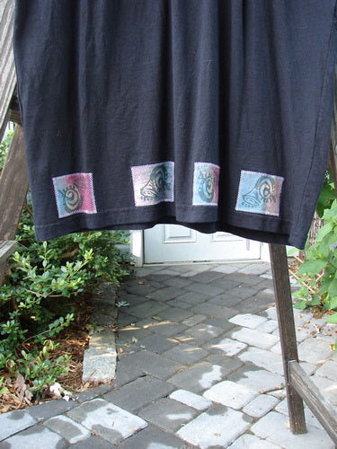 1992 Patched Little Storma Dress Music Black Small OSFA displayed on a wooden ladder, showcasing music-themed patches, deep rounded neckline, tapered lower, and gathered back with slits for hand-dyed silk ribbon.
