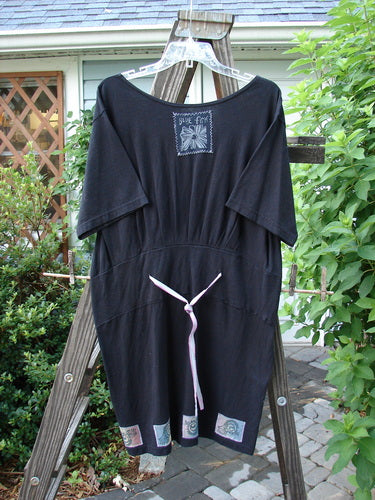 1992 Patched Little Storma Dress Music Black Small OSFA displayed on a wooden rack, featuring a deep rounded neckline, tapering lower, gathered top half rear, and vintage music-themed patches with variegated silk ribbon.