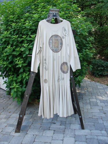 1994 Column Dress Belief Mist Size 1 displayed on a wooden stand outdoors, showcasing its full front button design, V-shaped neckline, longer sleeves, and abundant elongating paint in a spiritual theme.