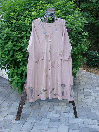 1993 Modernismo Duster Dress Columns Ash Pink OSFA displayed on a rack, showcasing vintage wooden buttons, front pockets, a scooped neckline, and a sweeping lower hem with a column and leaf pattern.