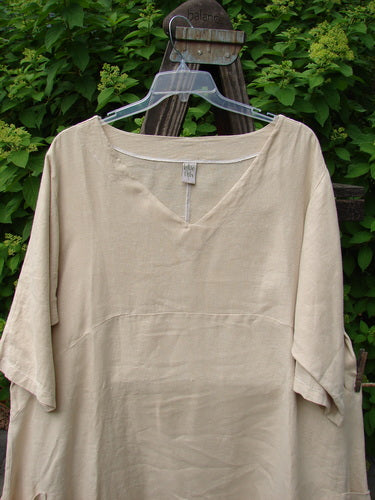 Barclay Linen Urchin Side Pocket Tunic Dress Unpainted Dried Wheat Size 2 displayed on a wooden stand, featuring a wide V-neckline, three-quarter sleeves, empire waist seam, and two exterior drop pockets.