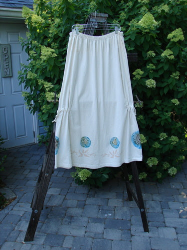 1999 Tie In Skirt Leaf Medallion Natural Size 2: A white skirt with blue trim hanging on a wooden rack, featuring an elastic waistband, draw cords, and detailed leaf medallion panels.