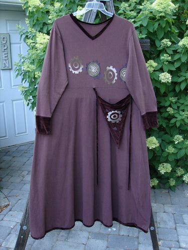 A 1996 Velvet Ornamental Pocket Dress Spirograph Spicewood Size 1. A purple dress with a design on it, featuring a velvet-accented neck, hem, and sleeve trim. The dress has a versatile tie-on, tie-off long velvet rippie cord. The image shows a close-up of a purple scarf and a plant.