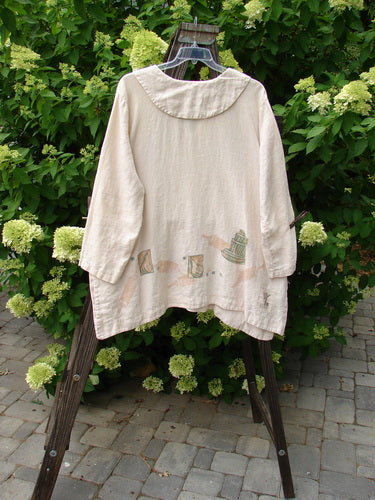 1999 Linen Antique Top Baker Tea Dye Size 1 displayed on a hanger, showcasing its V neckline, rounded drop collar, and hidden side pockets amidst an outdoor garden setting.