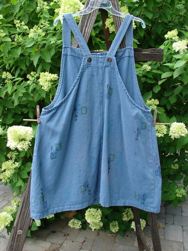 Barclay Canvas Urban Pocket Apron Jumper Mosaic Chair Blue Jean Size 1: A blue overalls with tear button accents and drop front pockets on a wooden rack.