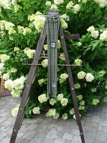 Image alt text: "Barclay Patched Scarf with linear green olive plaid on a wooden ladder with a cloth on it, outdoor garden vibe"